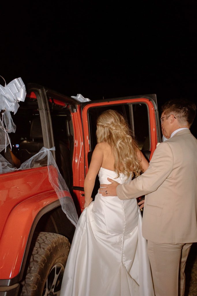 Groom helps bride into red jeep wrangler leaving their texas wedding reception.