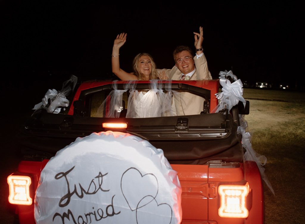 Bride and groom wave bye in red Jeep Wrangler after texas wedding reception.