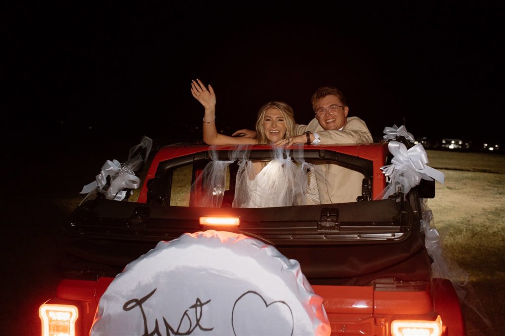 Bride and groom laugh and wave bye in red Jeep Wrangler after texas wedding reception.