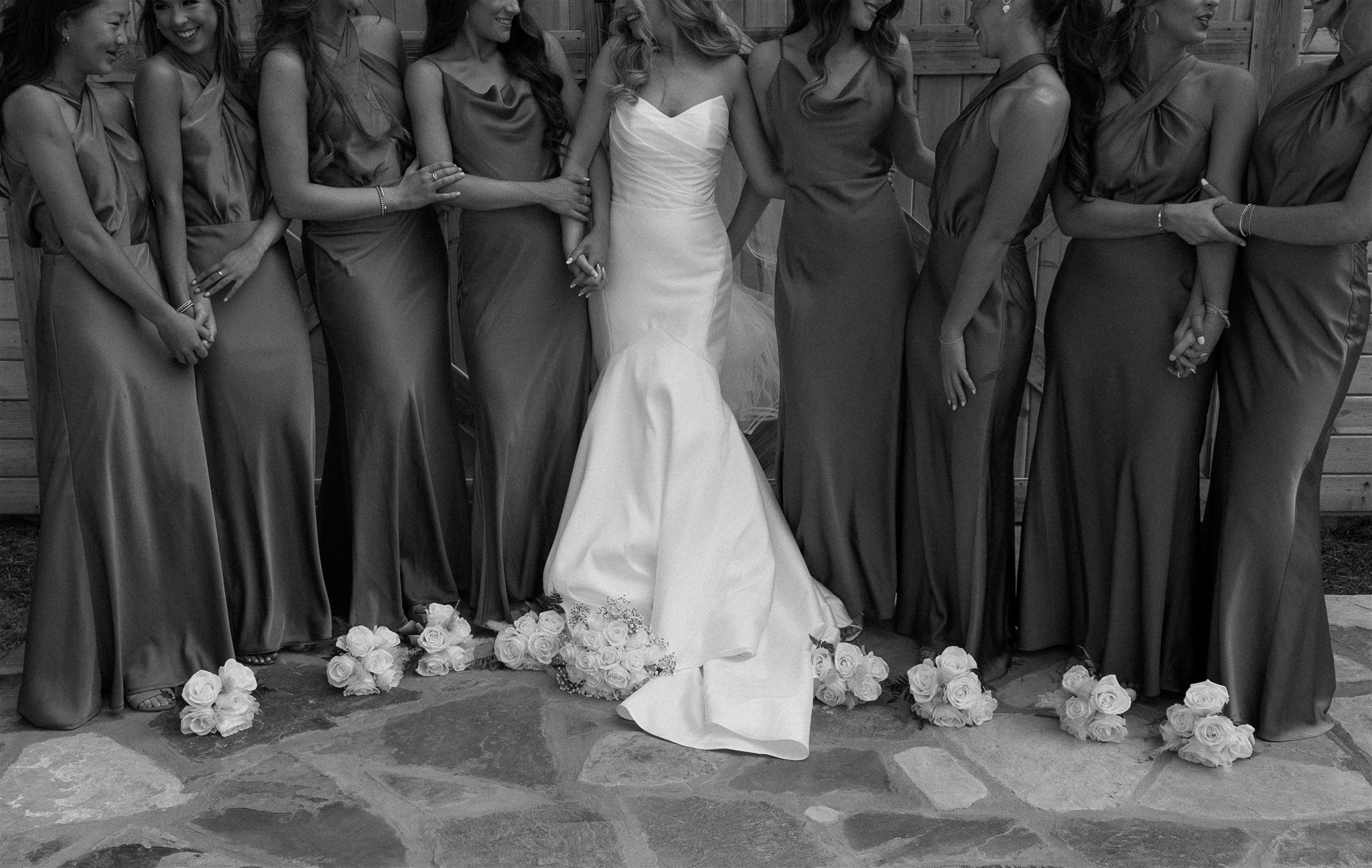 Bride and bridemaids candidly laugh with bouquets on the ground at their feet in black and white.