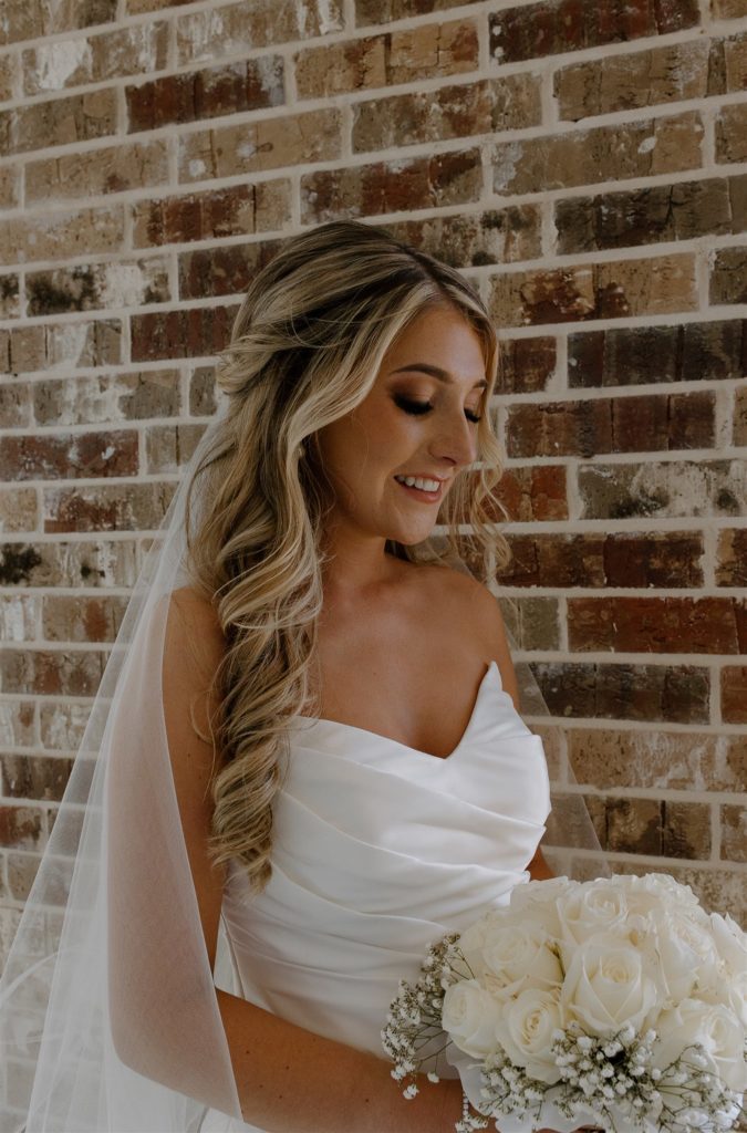 Bride smiles at white rose bouquet in front of brick wall.