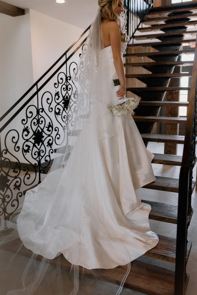 Bride stands on grand staircase holding bouquet during Texas wedding.